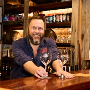 Hadyn Black holding two wine glasses at the bar in the Black and Ginger Wine Shed, serving a customer
