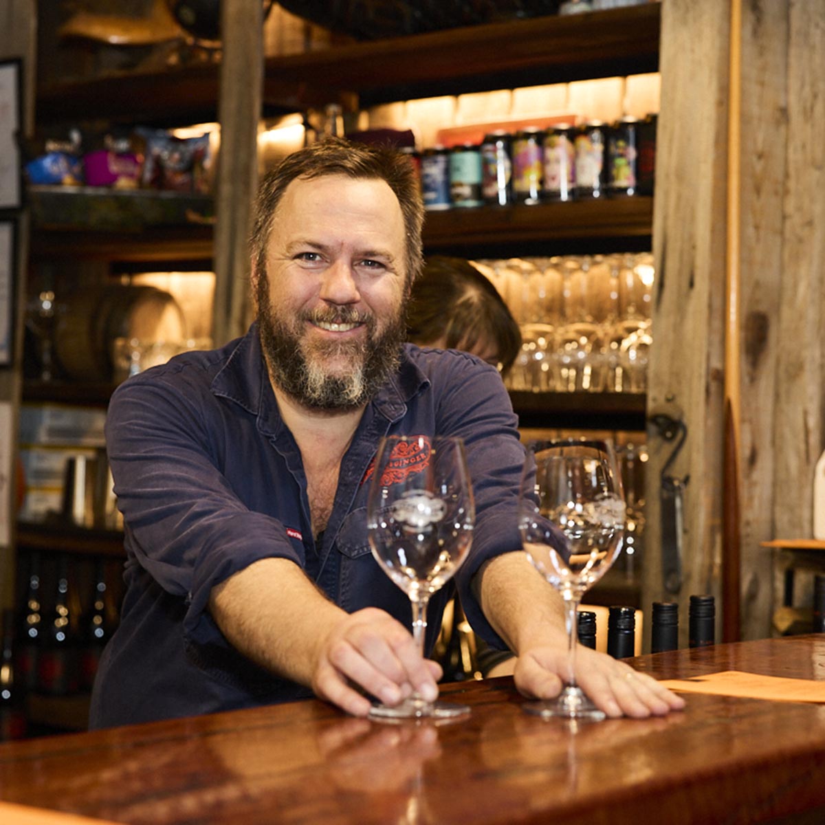 Hadyn Black holding two wine glasses at the bar in the Black and Ginger Wine Shed, serving a customer